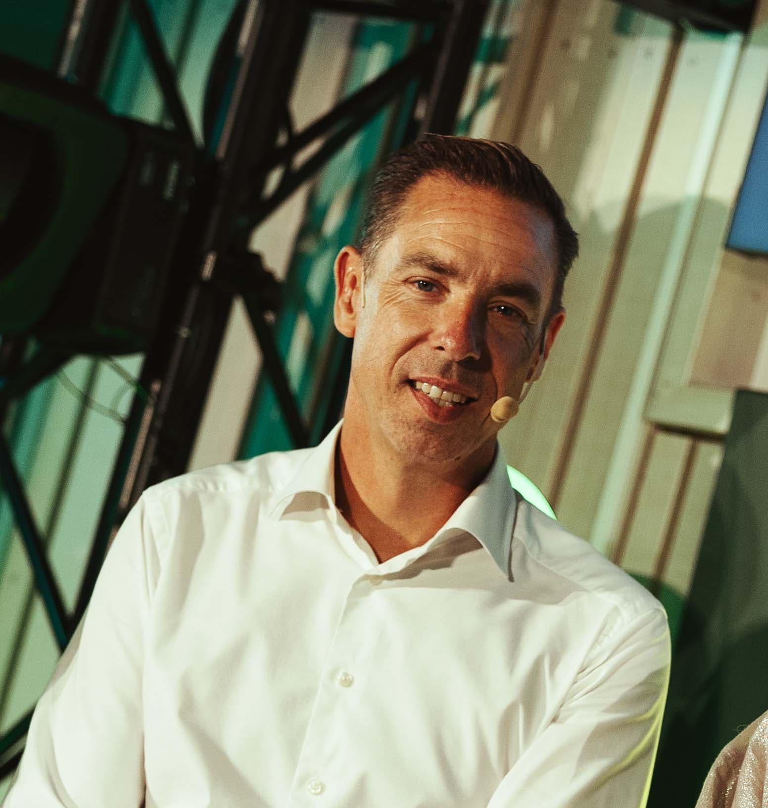 Man in a white shirt with a headset microphone, smiling against a backdrop of metal framework and a green-lit area.