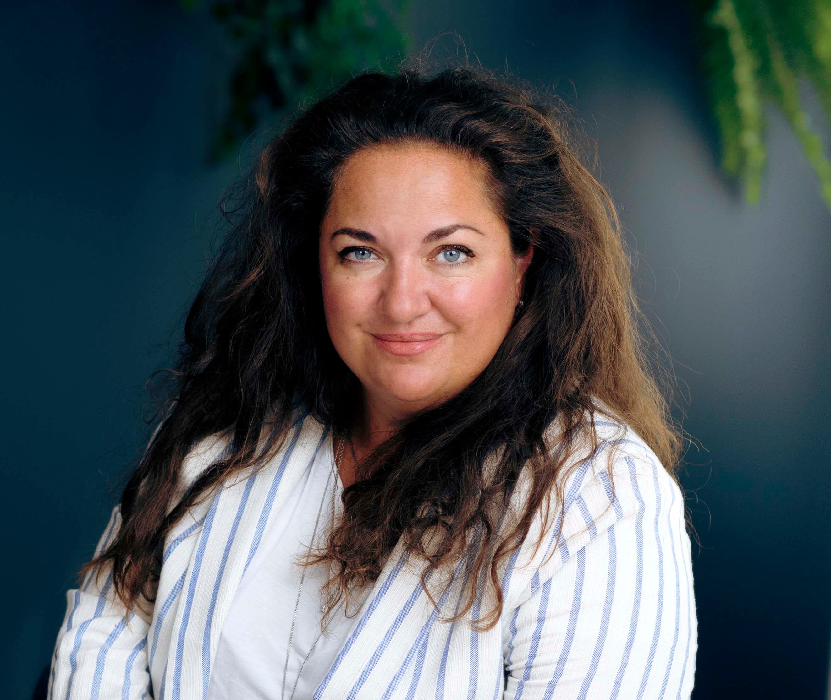Woman with long dark hair in a striped blazer, smiling softly, against a dark backdrop with green foliage.