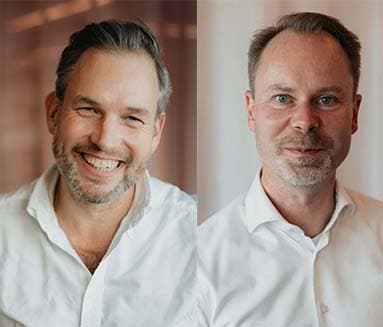 Two men in white shirts smiling, side by side against a softly lit background.