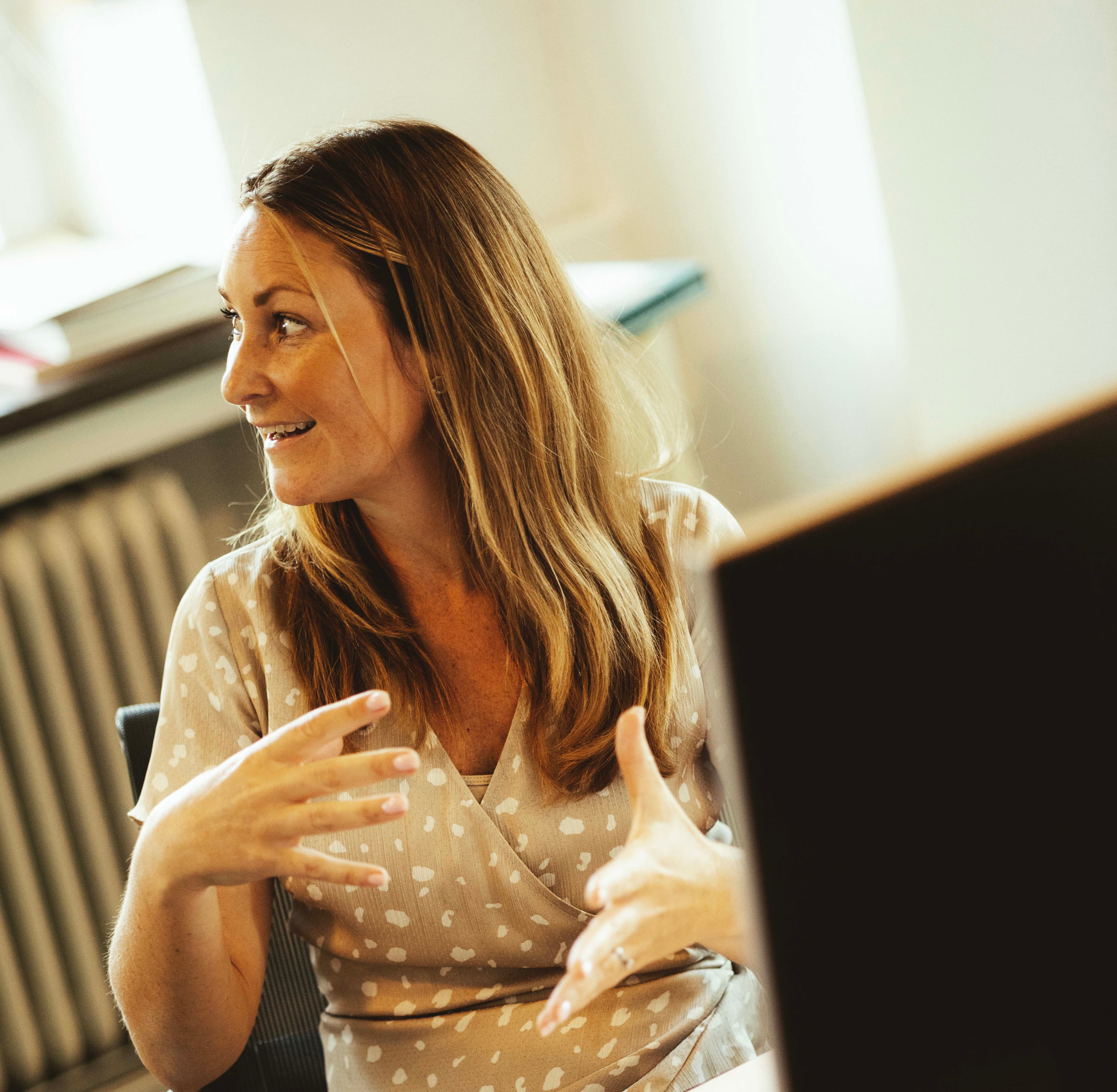 Woman with long hair gesturing while talking, seated indoors near a window with soft lighting and part of a computer screen visible in the foreground.
